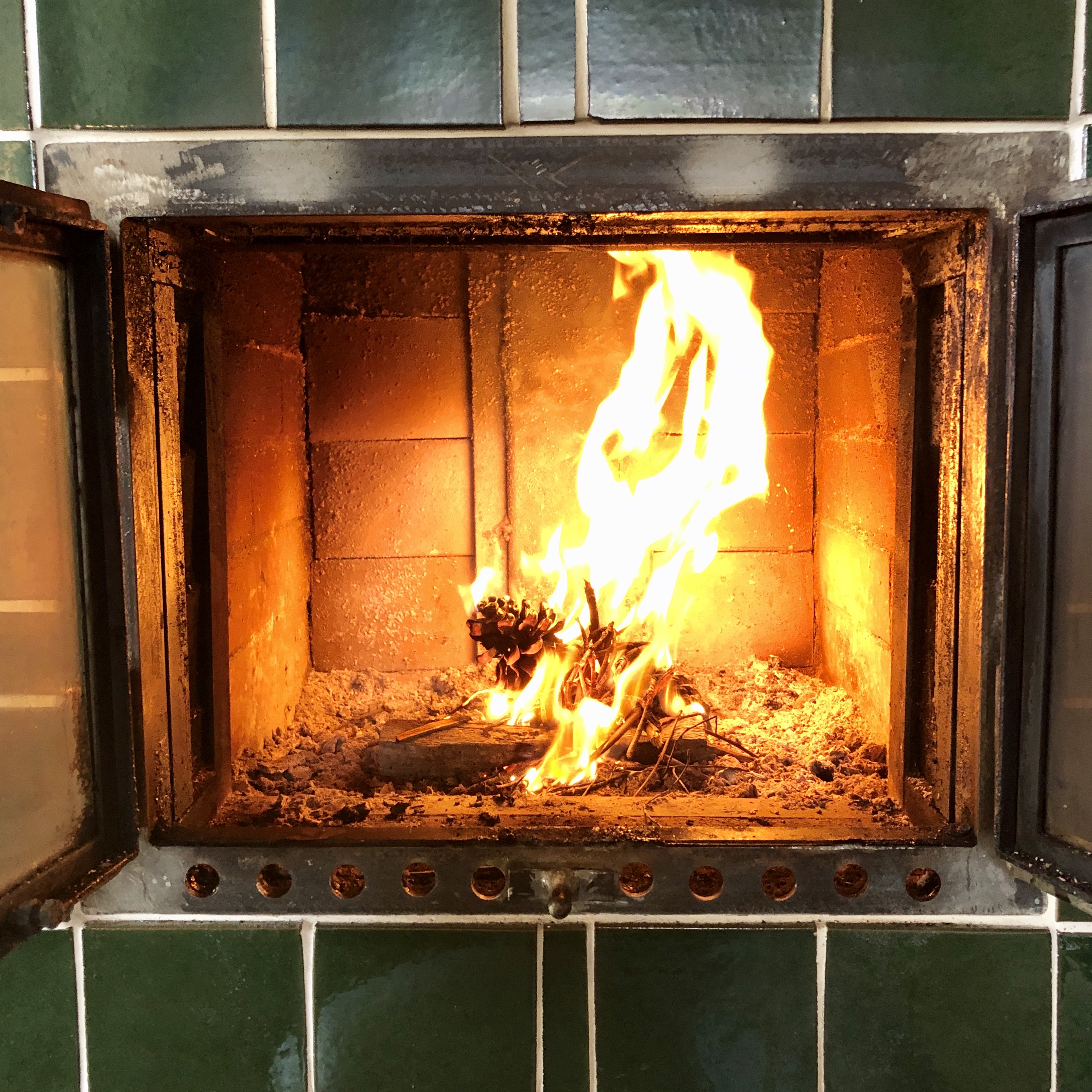 A log stove set into green-tiled hearth in Aljezur, catching flame with a single pine cone and dry kindling.